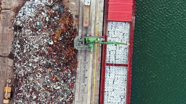 A large pile of scrap metal sits next to a cargo ship at the port. A crane is moving materials from the pile to the ship's open hold. Water is visible nearby.