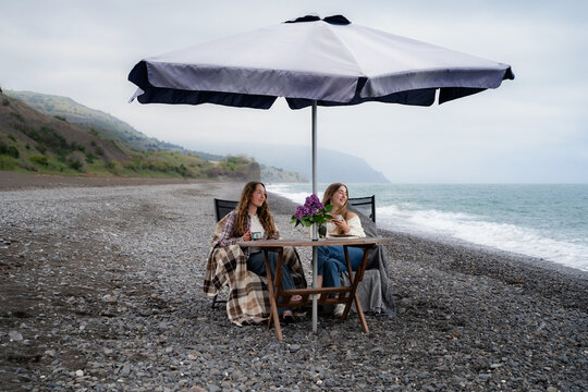 Two peaceful female friends are relaxing by the sea at a cozy seaside table during cloudy weather.
