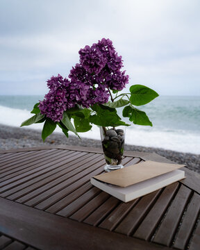 A lilac bouquet and a book on the seaside table, with an ocean view, in overcast weather.
