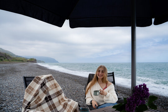 A relaxed, cute woman is enjoying a warm drink on the seaside, with an oceanic backdrop. She looks into the camera and smiles.