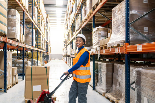 Young woman worker smiling while pushing pallet jack with goods in a large distribution warehouse