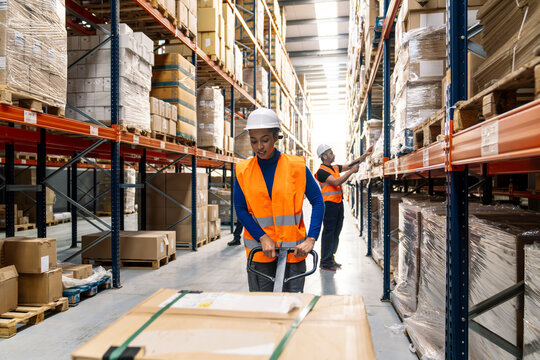 Female worker pulling pallet jack with boxes, colleague checking inventory, standing in busy warehouse aisles filled with goods