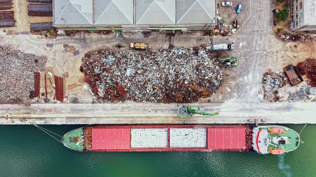A cargo ship is docked alongside a scrapyard. The ship is taking on materials from the nearby pile of scrap metal. Trucks and machinery are present at the site.