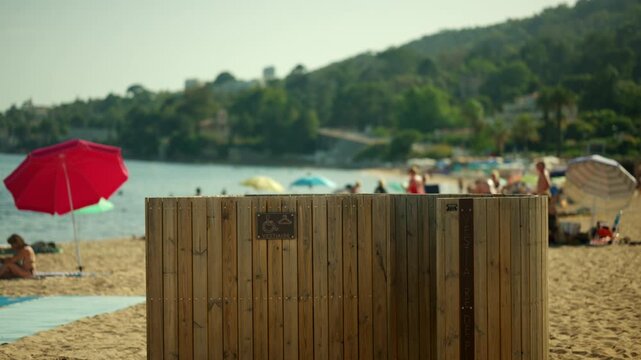 Wooden changing stall providing privacy for beach visitors, with a red umbrella and people relaxing by the sea on a sunny day