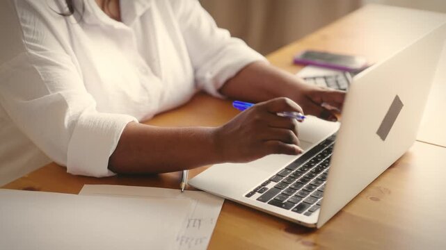 African woman working in office, closeup view of female hands on laptop keyboard. Workplace at home, accountant checking report in computer and making note in paper, tech support and customer service
