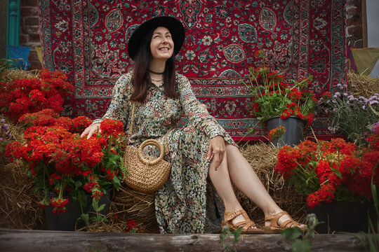 woman sits on stack of hay surrounded by bright flowers. She wears long dress and hat. backdrop features colorful pattern, adding to market setting