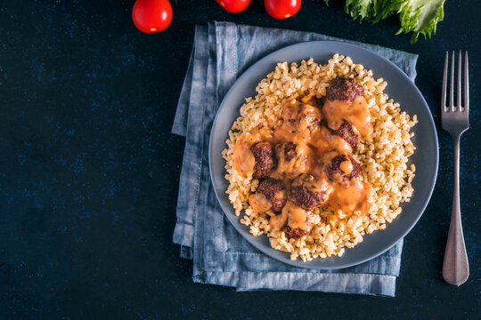 Delicious hot meatballs covered in savory gravy sauce served with bulgur or rice on a plate. Top view flat lay composition on dark textured background with copy space. Perfect for restaurant menu