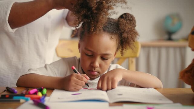Black schoolgirl doing home exercise in workbook, careful mother doing hairstyle. Morning in happy african american family before school, portrait of pretty mixed race girl holding pen and drawing