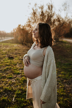 Smiling expectant mother holding baby bump outdoors at golden hour