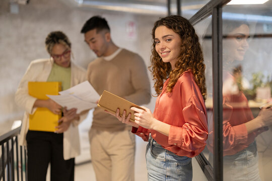 Smiling young businesswoman standing with diary near colleagues discussing over documents in illuminated corridor