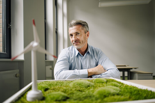 Smiling businessman sitting by wind turbine toy and moss frame on table at office