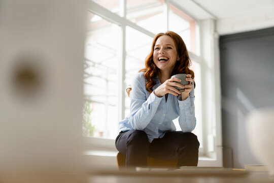 Portrait of laughing redheaded woman with tea bowl sitting on backrest in a loft