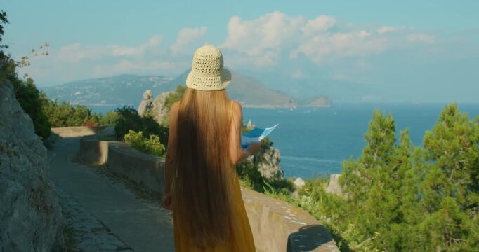 Woman walking a seaside path with a map overlooking blue bay and distant hills in summer