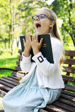 Excited Young Girl with Book on Park Bench in Sunny Garden