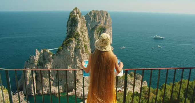Woman with map studies view of Faraglioni rocks from Capri cliff lookout in summer