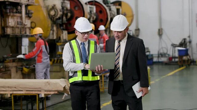 Engineers wearing hardhats review data on a laptop inside a busy factory, with heavy machinery and additional workers operating equipment in the background.