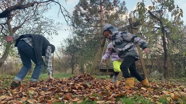 Young dad with daughter and son of primary school age in warm autumn clothes enjoying quality time together in the yard among fallen golden leaves. Cozy family autumn concept. The beauty of fall .