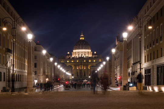 Symmetrical night view of the illuminated St. Peter's Basilica along Via della Conciliazione with motion blurred people in Rome, Italy