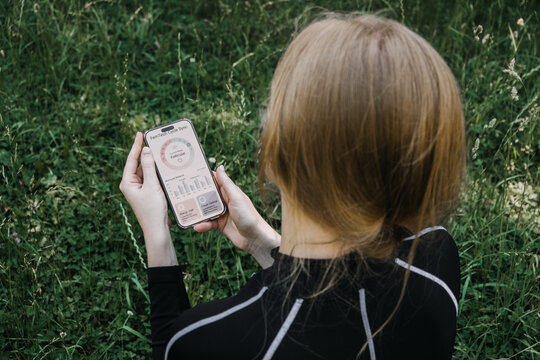 Woman sits on grass holding smartphone displaying hormone tracking application. cycle syncing workout, menstrual cycle app, femtech tracking, women health technology