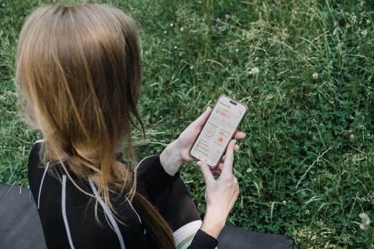 Woman sitting outdoors holding smartphone displaying fitness dashboard with heart rate and calorie metrics. AI fitness tracker data, smartwatch monitoring, biohacking lifestyle, urban wellness, heart
