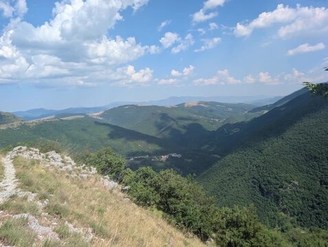 Pendio roccioso ed erboso che scende verso una valle circondata da colline verdi e crinali sovrapposti, illuminati da una luce morbida sotto un cielo parzialmente nuvoloso