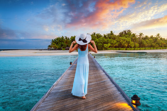 A beautiful woman walks down a pier over turquoise ocean towards a tropical island in the Maldives during sunset time