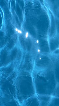 Close-up view of bright sun caustics and light patterns dancing on the bottom of a clear blue swimming pool with gentle ripples on the water surface, 4k