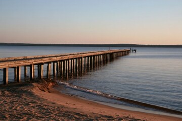 Fototapeta premium Long wooden pier extending over calm water at sunset with sandy beach foreground