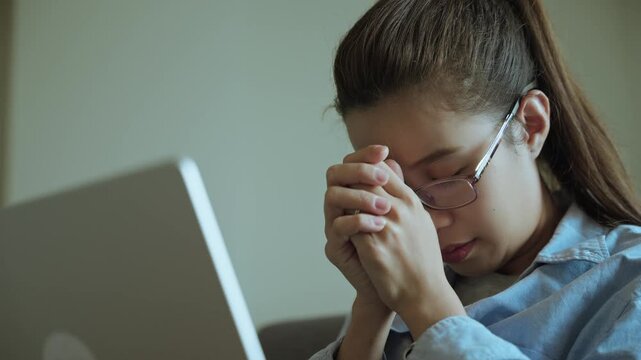 Stressed Young Asian Woman Closing Eyes and Praying While Working on Laptop at Home
