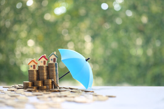 Miniature houses on stacked gold coins protected by a blue umbrella, concept of real estate insurance, property protection and financial security for home investment.