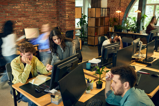 Tired employees working at computer desks while coworkers move through open office. Concept of workplace burnout prevention, workload balancing and employee wellbeing.