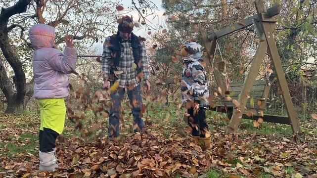 Young dad with daughter and son of primary school age in warm autumn clothes enjoying quality time together in the yard among fallen golden leaves. Cozy family autumn concept. The beauty of fall .