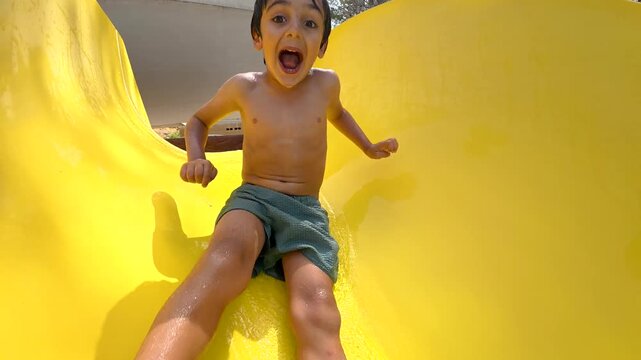 Young boy with a happy and surprised expression sliding down a yellow chute into a swimming pool, enjoying a sunny day at the waterpark during his summer vacation