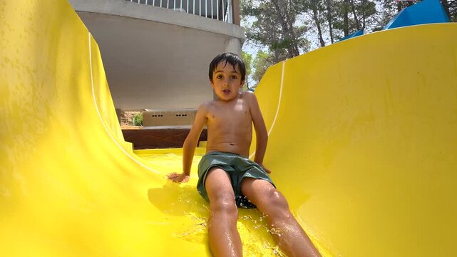 Exuberant little boy enjoying a sunny day at the water park, happily sliding down a bright yellow chute and splashing into the pool with a big smile, capturing the essence of summer fun