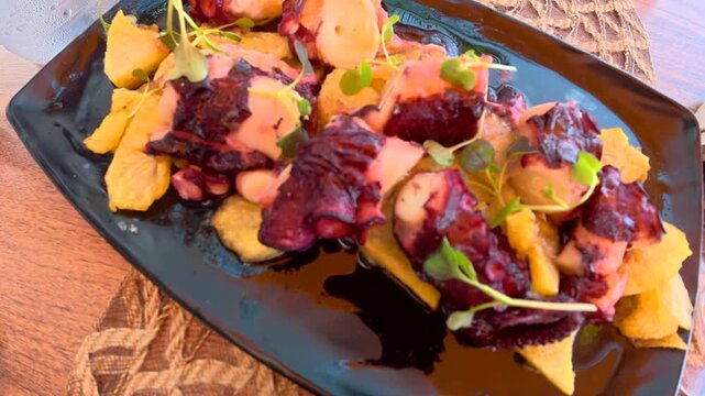 Close up view of a traditional spanish tapa of galician style octopus with potatoes and microgreens, a delicious seafood appetizer served in a black dish on a restaurant table during summer