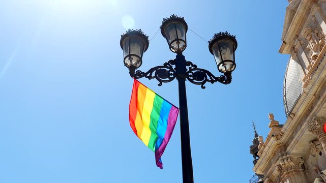 Brightly colored lgbtq pride flag waving on an ornate street lamp against a clear blue sky in the historic city of cartagena, spain, symbolizing freedom, love, and community celebration