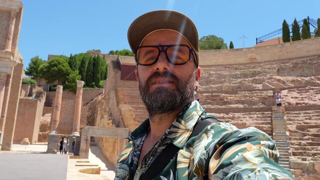 Young man vlogging with a selfie stick and talking about his trip while visiting the ancient roman amphitheater ruins in cartagena, a famous tourist destination in the murcia region of spain
