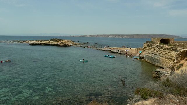 People enjoying a beautiful summer day on the rocky coast of tabarca island, spain, with clear turquoise water of the mediterranean sea and santa pola visible in the distant background