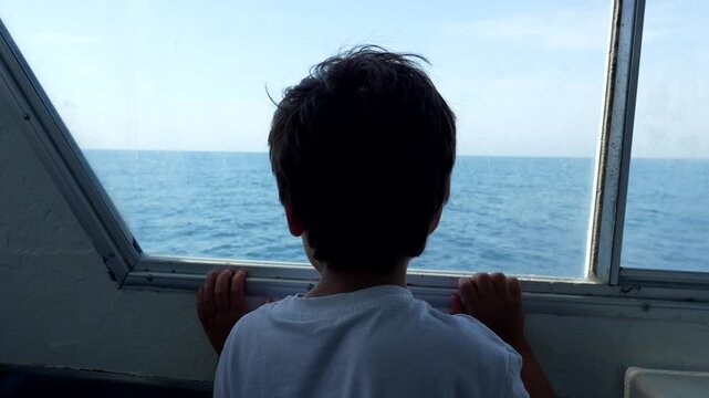 Back view of a young boy traveling by boat and looking out the window at the blue ocean during a sunny summer day trip to tabarca island in alicante, spain, representing childhood dreams and travel