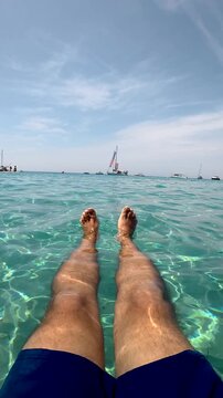 Point of view of a man's legs floating in calm, transparent turquoise sea water on a sunny day in tabarca island, spain, with sailboats on the horizon creating a serene vacation scene