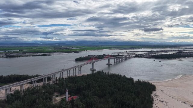 Dramatic aerial perspective captures the Thuan An Bridge's cable-stayed tower rising above the water, with the red deck stretching across the wide lagoon.
