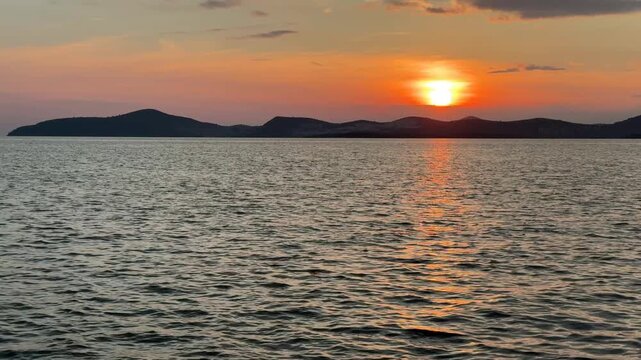 A calm, warm sunset over the Aegean Sea in Gulluk, Turkey. The sun hangs low in a hazy orange sky, casting a bright golden light path across the gently rippling water toward distant dark hills.
