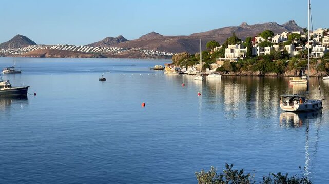 Scenic view of the calm, blue bay in Bitez, Turkey, with luxury boats anchored on the still water near a hillside resort town under a clear morning sky.
