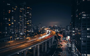 Fototapeta premium A bustling cityscape at night showcases elevated highways filled with light trails from speeding vehicles surrounded by towering buildings and illuminated streets