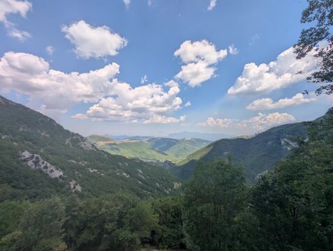 Vista ampia su pendii verdi e vallate profonde illuminate da luce naturale, con montagne che sfumano all&rsquo;orizzonte sotto un cielo parzialmente nuvoloso