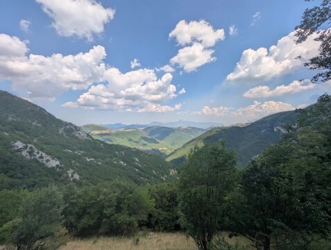 Vista ampia su pendii verdi e vallate profonde illuminate da luce naturale, con montagne che sfumano all&rsquo;orizzonte sotto un cielo parzialmente nuvoloso