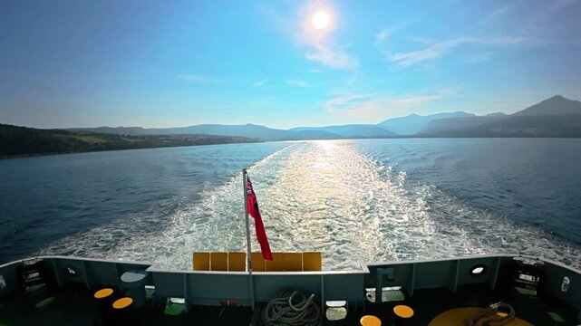 Ship stern view with flag waving in wind creating wake on water. Troon to Brodick ferry Scotland

View from the stern of a boat showing the water wake and a waving flag against a sunny ocean