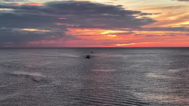 Wide aerial view shows multiple fishing boats dispersed across a vast ocean surface during early morning, with soft pink and orange sunrise colors painting the sky.