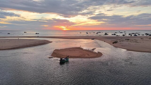 Aerial drone view of a sandy spit dividing river and ocean waters, with colorful fishing boats anchored in the sheltered estuary under a dramatic sunrise sky.