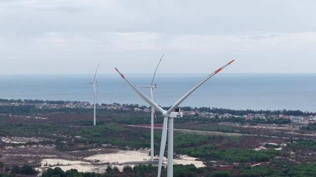 Dramatic close-up aerial shot orbiting a wind turbine with red-tipped blades spinning above the coastal landscape, the sea stretching to the horizon behind.